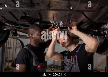 two interracial car mechanics in uniform checking car in automobile service with lifted vehicle, african and caucasian men repair a car together Stock Photo