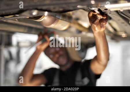 young african male car mechanic in uniform checking car in automobile service with lifted vehicle, focus on hands. Stock Photo