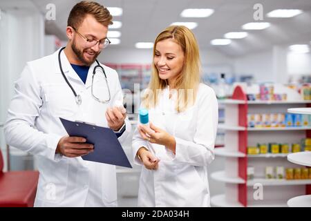 two young caucasian pharmacists are discussing medication, using a digital tablet and documents. man and woman in white medical gown working in pharmacy Stock Photo