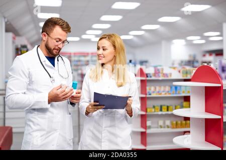 two young caucasian pharmacists are discussing medication, using a digital tablet and documents. man and woman in white medical gown working in pharmacy Stock Photo