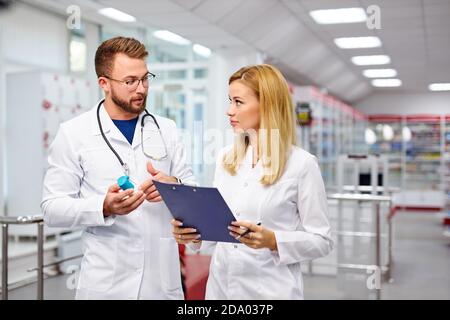 two young caucasian pharmacists are discussing medication, using a digital tablet and documents. man and woman in white medical gown working in pharmacy Stock Photo