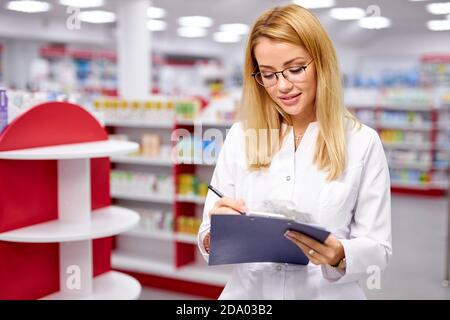 blonde druggist woman in uniform checking the assortment stock in pharmacy, read indications Stock Photo