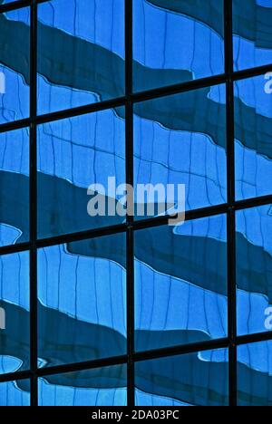 Cladding sheathing one of the high-rise office towers in Central District or Chung Wan, the heart of global business activities in Hong Kong, China, distorts the reflection of another corporate headquarters nearby with rippling blue and grey bands within a rigid black grid. Stock Photo
