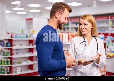 young beautiful caucasian pharmacist is working with customer in modern drugstore, serve the client, wearing white medical gown Stock Photo