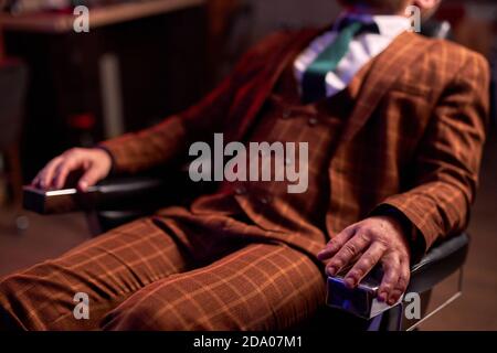 cropped male in stylish suit or tuxedo sit having rest in barber shop, unrecognizable business man enjoy cutting his beard Stock Photo