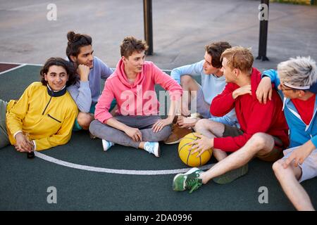 young friends sitting on basketball court, relaxing and taking break after game. talk, laugh, tell stories and jokes. youth concept Stock Photo