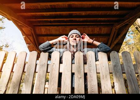 Portrait from below of a beautiful woman in the autumn season touching her hat leaning against the fence of a forest hut with orange and yellow tree l Stock Photo