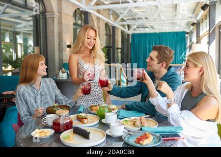blonde caucasian woman say a toast in honor of the birthday of friend or colleague in restaurant, they drink beverages and eat delicious tasty food, meal Stock Photo