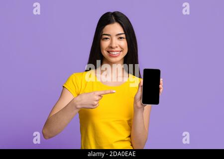 Check This. Cheerful Asian Lady Pointing At Smartphone With Black Blank Screen Stock Photo