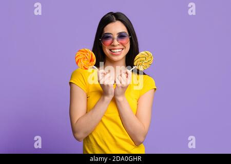Sweet Treat. Pretty young summer lady covering eye with big bright lollipop and looking at camera, yellow background with copy space Stock Photo