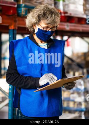 Side view focused female worker in blue vest and face mask taking notes in clipboard while standing near stacks in warehouse Stock Photo