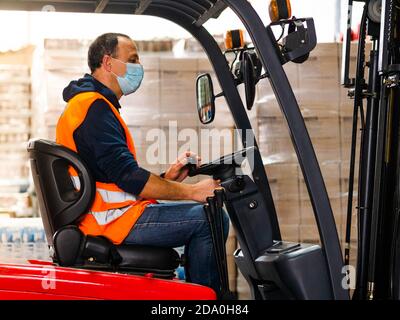 Side view unrecognizable male worker in high visibility vest and face mask driving autoloader machine while working in warehouse Stock Photo