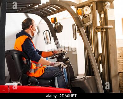 Side view unrecognizable male worker in high visibility vest and face mask driving autoloader machine while working in warehouse Stock Photo