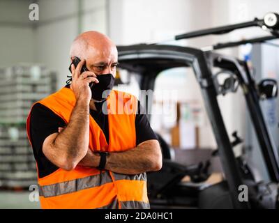 Serious adult male manager in uniform and protective mask standing with arms crossed near warehouse autoloader and having conversation on mobile phone Stock Photo