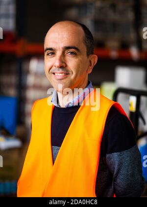 Happy adult male worker wearing orange high visibility vest standing near warehouse racks and looking at camera Stock Photo