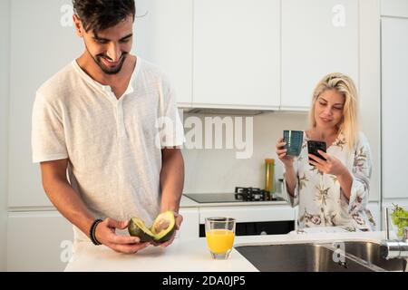 man with fresh avocado and orange juice in glass standing in kitchen with Woman using smartphone and drinking coffee at breakfast Stock Photo