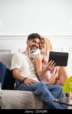 Happy young couple cuddling and browsing tablet while sitting together on cozy sofa near table in the morning Stock Photo
