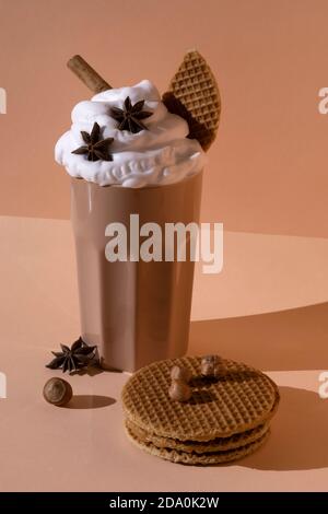 Glass with chocolate cocktail with whipped cream garnished with star anise and cinnamon stick placed on table with hazelnuts and cookies on pastel bro Stock Photo