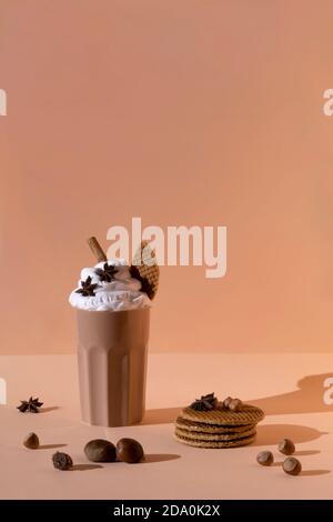 Glass with chocolate cocktail with whipped cream garnished with star anise and cinnamon stick placed on table with hazelnuts and cookies on pastel bro Stock Photo
