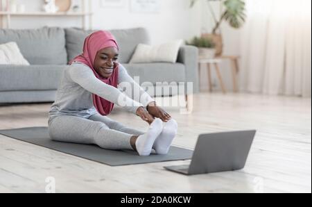 Black muslim woman doing stretching exercises in front of laptop at home Stock Photo