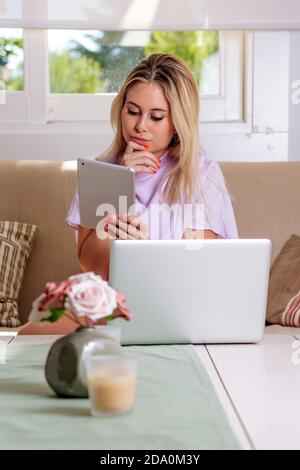 Thoughtful female freelancer sitting on sofa in living room and browsing on tablet near netbook while working on remote project Stock Photo
