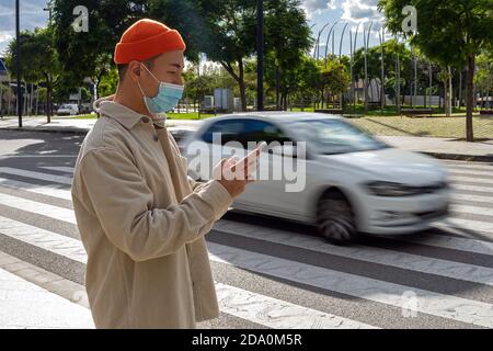 Side view focused young Asian male wearing casual outfit and orange hat browsing mobile phone while standing on sunny street near moving car Stock Photo