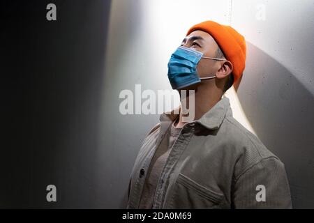 Concentrated Asian male in casual wear and protective face mask standing calmly in blurred gray room and looking away Stock Photo