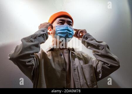 Concentrated Asian male in casual wear and protective face mask standing calmly in blurred gray room and looking away Stock Photo