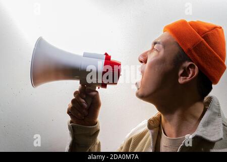 Side view of overwhelmed male protesting in casual clothes screaming in loudspeaker while standing against gray wall in dark room Stock Photo