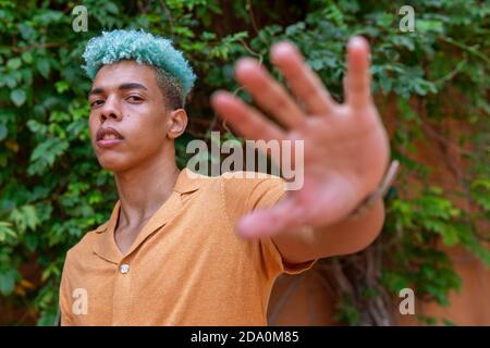 Confident African American young male hipster with blue curly hair reaching hand towards camera while standing on street Stock Photo