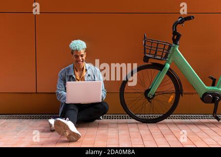 Smiling black male hipster with dyed hair using laptop while working on remote project on the street near modern bike Stock Photo