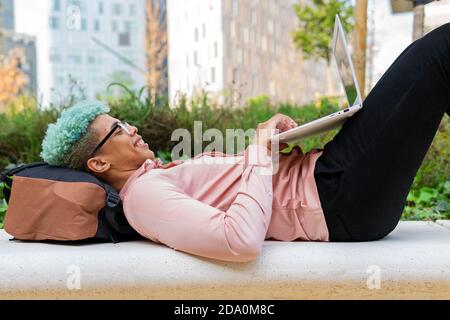 Side view of smiling African American male student with blue hair lying on backpack in university campus and typing on laptop while working on homewor Stock Photo