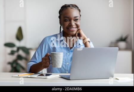 Pensive young black female freelancer drinking coffee at workplace in home office Stock Photo