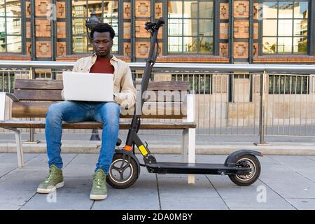 Concentrated African American male freelancer sitting on bench near electric scooter and working on project while using netbook in city Stock Photo