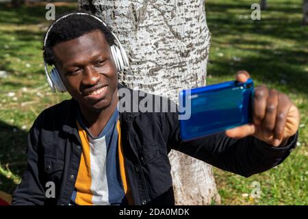 Handsome black male in wireless headphones sitting near tree in summer park and taking selfie on smartphone Stock Photo