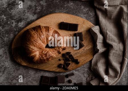 From above of delicious fresh crusty croissant and pieces of dark chocolate composed on wooden board on shabby table with linen fabric Stock Photo