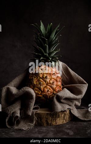 Still life composition with whole ripe pineapple wrapped in textile on wooden stand against black background Stock Photo