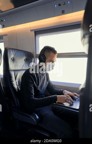 Young male in medical mask and headphones browsing data on laptop while riding modern train Stock Photo
