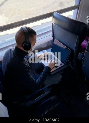 Young male in medical mask and headphones browsing data on laptop while riding modern train Stock Photo