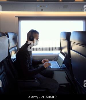Young male in medical mask and headphones browsing data on laptop while riding modern train Stock Photo