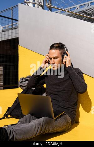 Serious young male entrepreneur wearing casual clothes using laptop and wireless headphones while working in park Stock Photo
