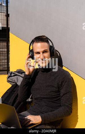 Serious young male entrepreneur wearing casual clothes using laptop and wireless headphones while working in park Stock Photo