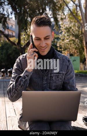 Full body of cheerful male freelancer sitting on bench and typing on laptop while talking on phone working outside Stock Photo