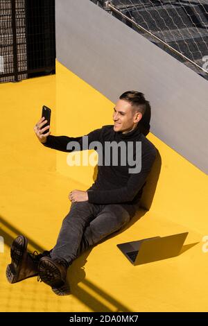 From above full length of happy young male remote employee having video conversation while sitting on yellow surface with laptop Stock Photo