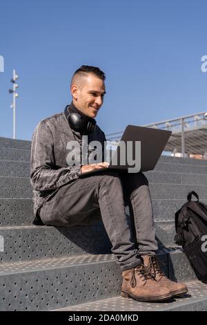 Cheerful male freelancer in headphones sitting on stairs and typing on laptop while working outside Stock Photo