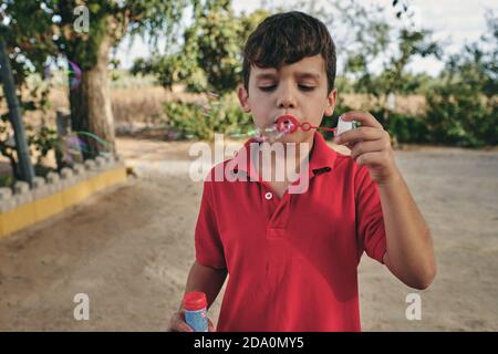 Adorable boy in summer outfit standing in park and blowing soap bubbles while having fun at weekend Stock Photo