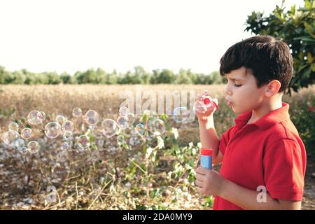 Adorable boy in summer outfit standing in park and blowing soap bubbles while having fun at weekend Stock Photo