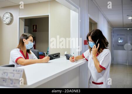 Concentrated female nurse wearing white medical suit and face mask taking notes in clinical record chart while standing near modern hospital reception Stock Photo