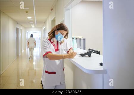 Concentrated female nurse wearing white medical suit and face mask taking notes in clinical record chart while standing near modern hospital reception Stock Photo