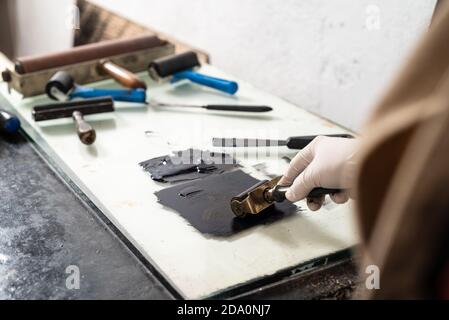 Closeup hand of crop anonymous artisan in glove using braver and spreading ink on sheet for engraving while working in professional workshop